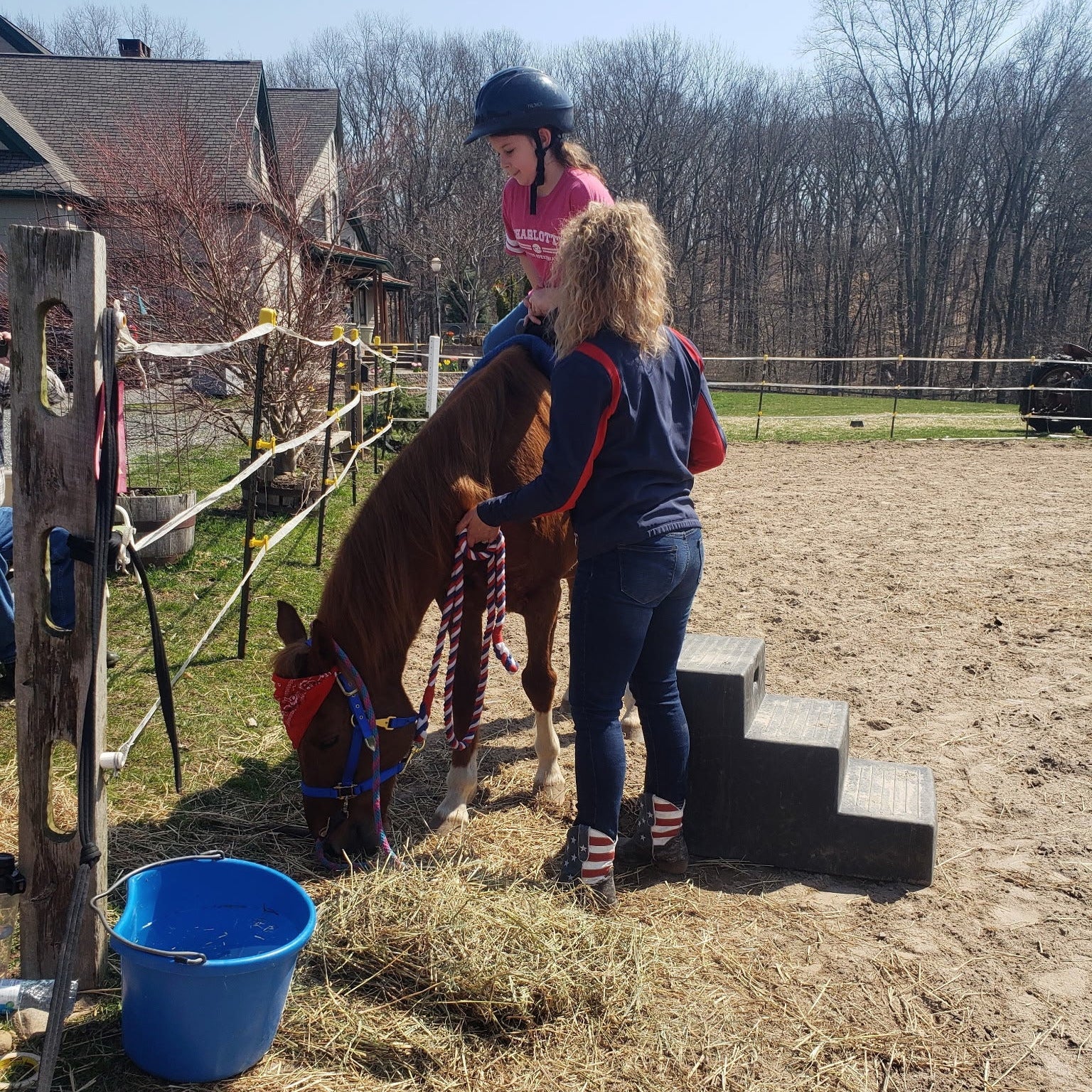 Woman helping a child ride a pony on a farm with a blue bucket nearby.